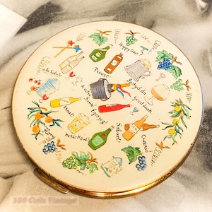 Könnte beinhalten: Runder Vintage-Kompakt-Make-up-Spiegel mit goldenem Rand und cremefarbener Oberfläche. Der Spiegel ist mit farbenfrohen Illustrationen von Flaschen, Gläsern und Sätzen wie "Happy Days" und "Proost" verziert. Das Design umfasst Trauben und Orangenäste.