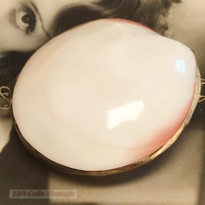 Könnte beinhalten: Eine ovale, vintage Schmuckschatulle in Muschelform mit weißem und rosa Innenraum. Sie hat einen goldfarbenen Metallrand und Scharniere. Der Text "100 Cells Vintage" ist unten sichtbar.