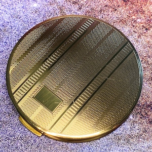 May include: A round gold-tone metal compact mirror featuring a textured surface with alternating vertical stripes of fine cross-hatching and smooth polished bands, including a small rectangular blank plaque centered on the front cover.