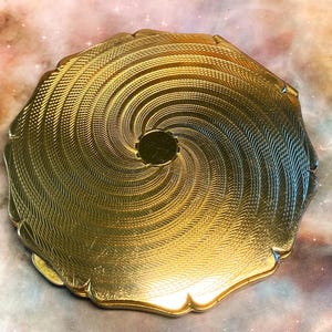 May include: A round gold-tone metal compact case featuring a scalloped edge and a textured surface with an intricate spiral pattern radiating from a smooth, circular center point. The metallic finish reflects light across the engraved design.