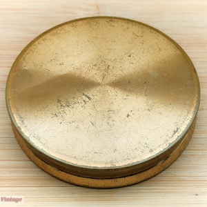 Könnte beinhalten: Ein runder, goldfarbener Metalldeckel mit leicht strukturierter Oberfläche. Der Deckel hat ein abgenutztes, gealtertes Aussehen.