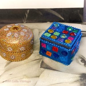 Könnte beinhalten: Zwei dekorative Boxen. Eine ist rund, goldfarben und mit Glitzer und Strasssteinen bedeckt. Die andere ist quadratisch, blau und mit bunten Edelsteinen verziert. Beide Boxen sind geschlossen und scheinen Schmuck- oder Schmuckkästchen zu sein.