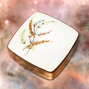 May include: A square metal compact case with a white enameled lid featuring a delicate hand-painted floral motif in shades of orange, green, and blue. The compact has a polished gold-tone metal frame and a hinged closure along the side edge.