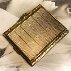 May include: A vintage gold-toned compact with a textured, grid-like pattern on the front. The edges are adorned with a delicate floral design. The compact is square and appears to be made of metal.