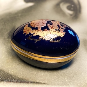 May include: A dark blue, egg-shaped trinket box with gold detailing. The top has a gold illustration and the words "Limoges Castel" in gold script. The box is edged with a gold band.