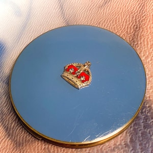 May include: A round, light blue compact mirror with a metallic gold-tone rim. Centred on the smooth, reflective light blue surface is a small, detailed emblem of a crown featuring red enamel accents and gold-tone metal detailing.