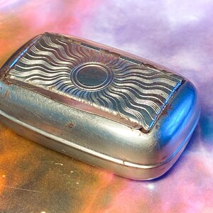May include: A rectangular silver-toned metal box with rounded corners. The lid features a central circular design surrounded by radiating wavy lines. The surface shows signs of wear and oxidation along the edges of the lid and the base.
