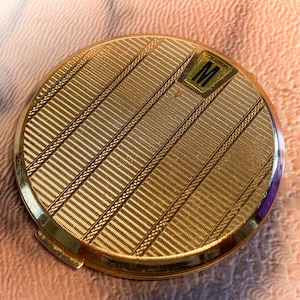 May include: A round gold-tone metal compact mirror featuring a textured surface with diagonal engraved lines and a small rectangular inset at the top right corner containing the black letter M. The compact has a polished finish and a hinged closure.
