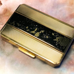 May include: A rectangular gold-tone metal case featuring a textured cross-hatch pattern on the top and bottom sections, with a central horizontal black band that displays visible wear and surface chipping across its dark finish.