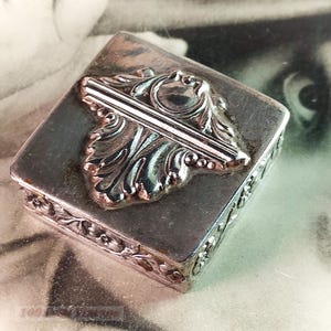 Può includere: Una piccola scatola quadrata argentata con dettagli floreali e a foglie. Il coperchio presenta un design a foglia stilizzata in rilievo con una barra centrale. I bordi della scatola sono decorati con motivi floreali.