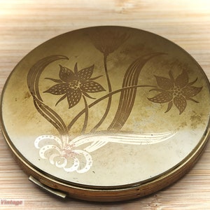 May include: A round, gold-colored compact with a floral design. The design features stylized flowers and leaves in a light gold color.