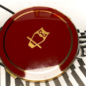 May include: A round, deep red compact mirror with a polished gold-tone metal rim. The centre of the lid features a gold-coloured graphic illustration of an owl perched on a branch. The compact has a smooth, glossy finish and a circular gold border.