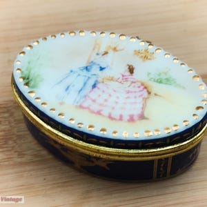May include: An oval porcelain trinket box with a blue and gold trim. The lid features a hand-painted scene of two women in colorful dresses.