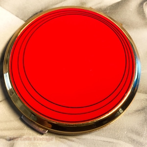 May include: A round, vintage compact with a bright red center and gold-toned trim. The red surface is accented with three concentric black lines. The compact has a small, silver-colored clasp. The background is a black and white image.