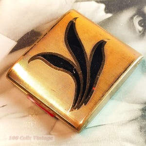 Op de afbeelding: Een vintage goudkleurige compact met een zwart bloemmotief. De vierkante compact heeft een getextureerd oppervlak en een rode accent. De achtergrond is een vintage foto. De tekst "100 Cents Vintage" is zichtbaar.