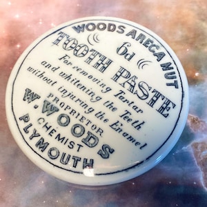 Könnte beinhalten: Ein runder weißer Keramikdeckel mit schwarzem Aufdruck: WOODS ARECANUT TOOTH PASTE, 6d, For removing Tartar and whitening the Teeth without injuring the Enamel, PROPRIETOR W. WOODS CHEMIST PLYMOUTH. Der Deckel hat eine glatte Oberfläche.
