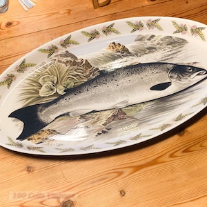 Könnte beinhalten: Eine ovale Servierplatte aus Keramik mit einer detaillierten Abbildung eines Lachses, der in einem Fluss schwimmt. Der Rand ist mit einem grünen Farnmuster verziert. Der Text SALMON Salmo Salar ist nahe dem Fisch auf cremefarbenem Grund.