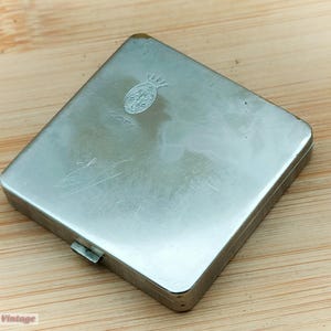 Może przedstawiać: Srebrne metalowe pudełko na papierosy z wypukłym wzorem ananasa i napisem "Coty" na górze.