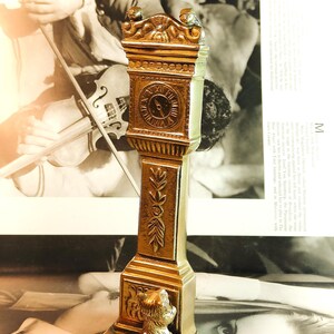 May include: A gold-colored miniature grandfather clock figurine. The clock face shows Roman numerals. The base features a small dog statue. The clock is set against a black and white background with text.