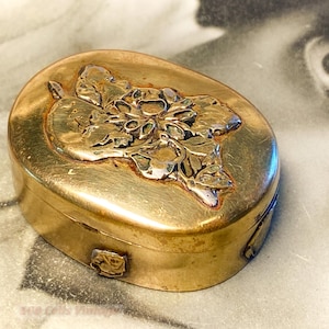 May include: An antique, oval-shaped gold-coloured metal box. The lid features a raised floral design in a lighter gold colour. The box has a hinged design and a small, decorative clasp. The background is a black and white photograph.