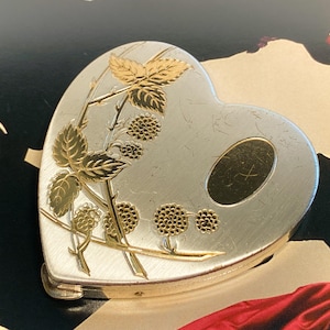 May include: A heart-shaped silver-tone metal compact case featuring an engraved gold-tone design of a blackberry branch with leaves and fruit. The surface includes a smooth, oval-shaped gold-tone plaque on the right side for potential engraving.