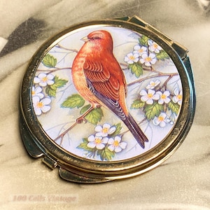 May include: A vintage compact mirror with a gold-tone frame. The mirror's face features a detailed illustration of a red bird perched on a branch with white flowers and green leaves. The compact is round.