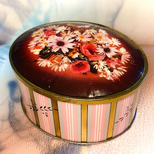 May include: An oval metal tin with a lid featuring a vibrant floral arrangement of white daisies and red poppies against a dark red background. The sides of the container are decorated with vertical stripes in pink, white, and gold, accented with small black leaf motifs.