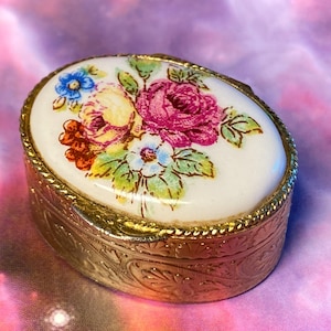 May include: An oval metal trinket box with a gold-toned finish and an embossed leaf pattern on the sides. The lid features a white ceramic inlay decorated with a colourful floral print of pink roses, yellow flowers, and blue blossoms with green leaves.