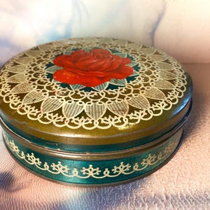 May include: A round metal tin featuring a dark green base colour with a prominent red rose illustration at the centre. The lid is decorated with a white lace-like pattern surrounding the rose, and the side of the tin has a white filigree border.