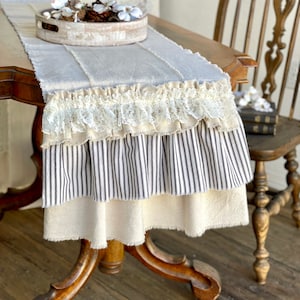 May include: A beige table runner with a white lace trim, black and white striped fabric, and a frayed edge. The runner is on a wooden table with a wooden chair in the background.