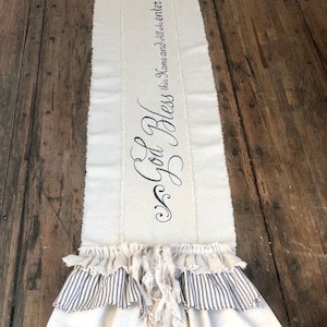 May include: A white table runner with a black inscription that reads "God Bless this Home and all who enter". The runner has a ruffled edge with a striped fabric underneath.