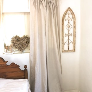 May include: A pair of off-white linen curtains with a simple tie-top design hang from a black metal rod. The curtains are gathered at the top and hang loosely, creating a relaxed and casual look. The curtains are in a bedroom setting with a wooden bed and a decorative metal window frame on the wall.