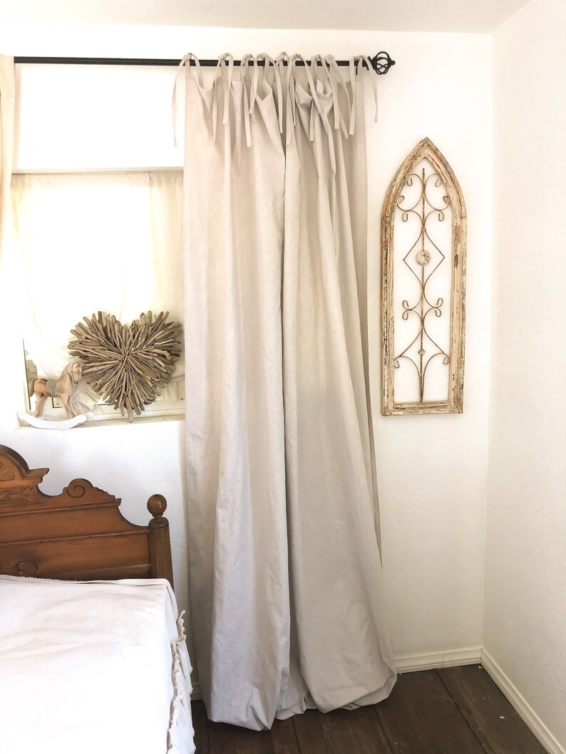 May include: A pair of off-white linen curtains with a simple tie-top design hang from a black metal rod. The curtains are gathered at the top and hang loosely, creating a relaxed and casual look. The curtains are in a bedroom setting with a wooden bed and a decorative metal window frame on the wall.