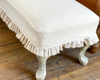 May include: A white linen bench with a ruffled edge and distressed white painted legs. The bench is sitting on a wooden floor.