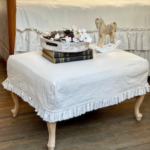May include: A white linen ottoman with a ruffled edge and wooden legs. The ottoman is decorated with a white tray, a stack of books, and a white rocking horse.