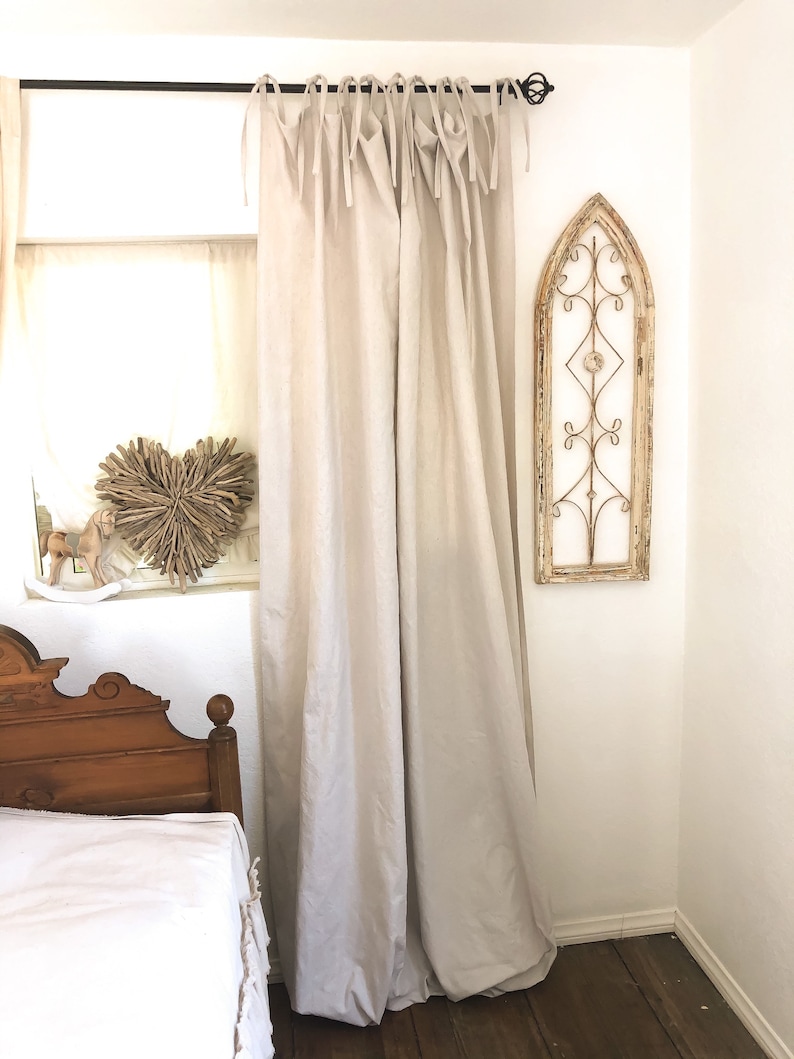 May include: A pair of off-white linen curtains with a simple tie-top design hang from a black metal rod. The curtains are gathered at the top and hang loosely, creating a relaxed and casual look. The curtains are in a bedroom setting with a wooden bed and a decorative metal window frame on the wall.