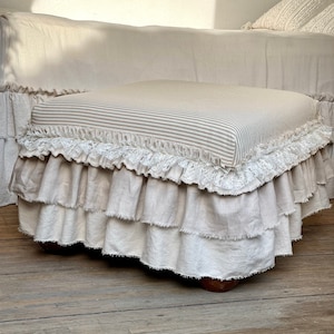 May include: A square ottoman with a striped beige and white fabric top and a layered ruffle skirt. The skirt is made of a natural linen fabric with a white lace trim.