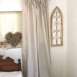 May include: A pair of light beige linen curtains with a simple gathered design and tied with fabric loops. The curtains are hanging from a black metal rod.
