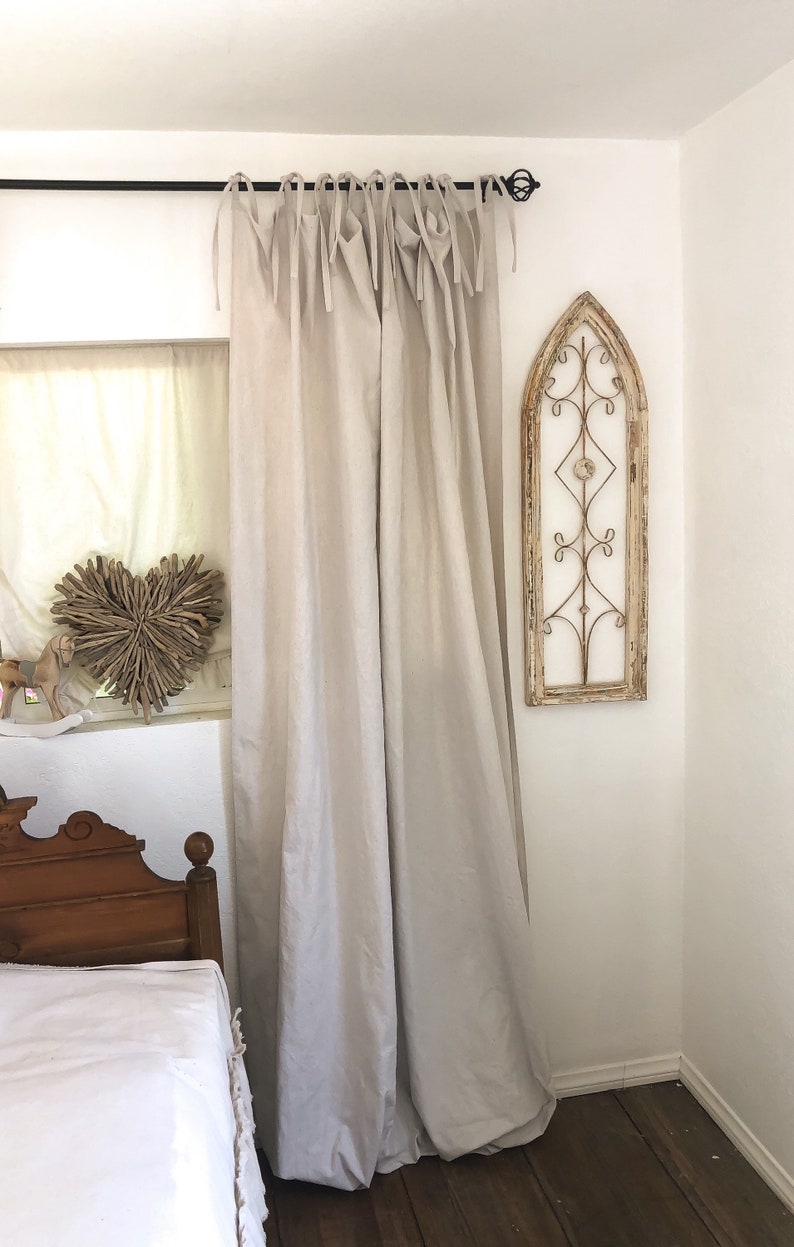 May include: A pair of light beige linen curtains with a simple gathered design and tied with fabric loops. The curtains are hanging from a black metal rod.