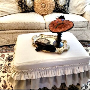 K&ouml;nnte beinhalten: Ein wei&szlig;er Ottoman aus Leinen mit einem R&uuml;schenrock. Der Ottoman steht auf einem gemusterten Teppich vor einem wei&szlig;en Sofa mit Kissen. Ein dekoratives Tablett mit einem Hut und Handschuhen befindet sich auf dem Ottoman.