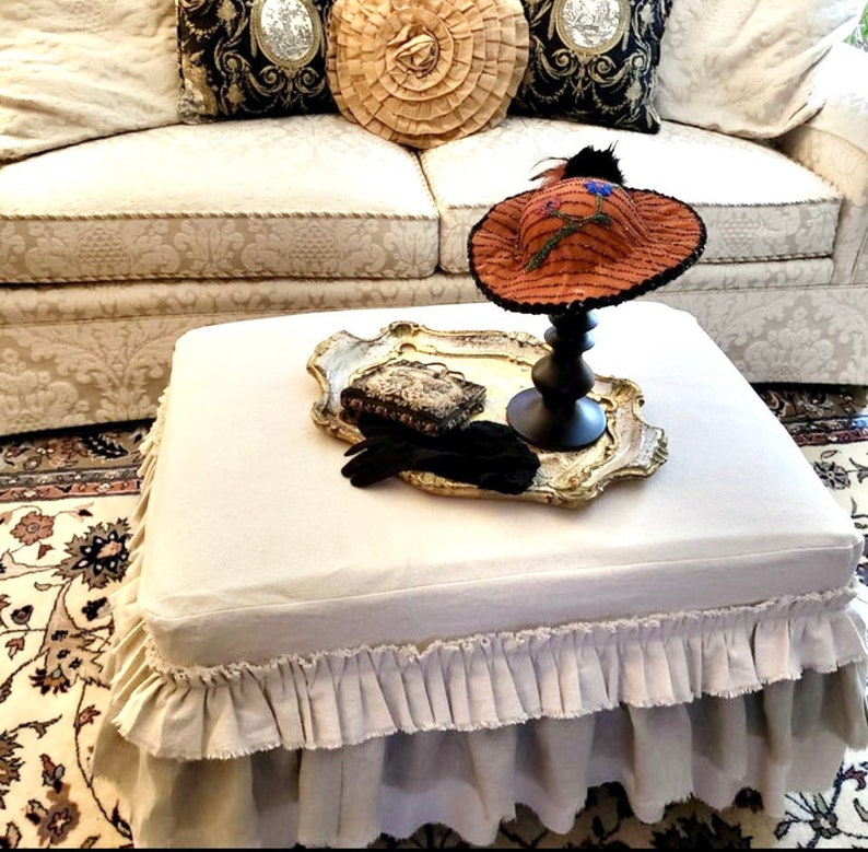K&ouml;nnte beinhalten: Ein wei&szlig;er Ottoman aus Leinen mit einem R&uuml;schenrock. Der Ottoman steht auf einem gemusterten Teppich vor einem wei&szlig;en Sofa mit Kissen. Ein dekoratives Tablett mit einem Hut und Handschuhen befindet sich auf dem Ottoman.