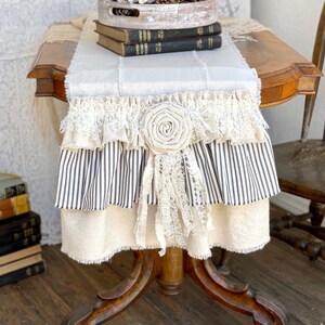 May include: A white and black striped table runner with a lace trim and a large white fabric flower. The runner is on a wooden table with a stack of books and a decorative tray with white flowers and pine cones.