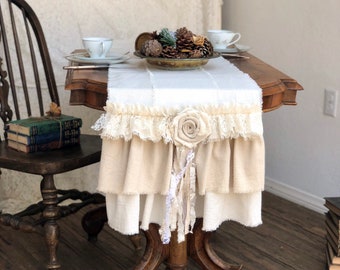 May include: A white linen table runner with a lace trim and a large white fabric flower in the center. The runner is on a wooden table with two teacups and a bowl of pine cones.