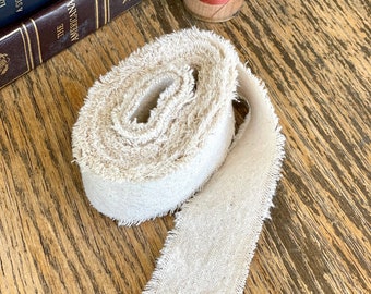 May include: A roll of natural-colored fabric trim with frayed edges. The trim is rolled up and lying on a wooden surface.