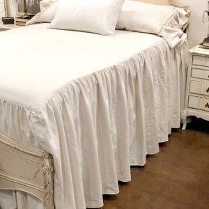 May include: White linen bed skirt with a ruffled edge. The skirt is on a white bed with white pillows. The bed has a white headboard with ornate carvings.