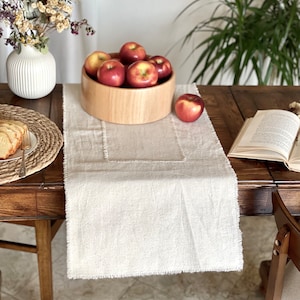 May include: A rustic wooden table setting featuring a beige linen table runner with a frayed edge. A wooden bowl filled with red apples sits atop the runner. A white vase with dried flowers and a plate of sliced bread complete the scene.