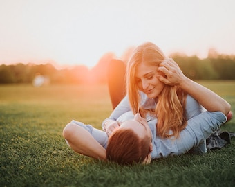 May include: Two individuals lying on green grass in a park. One person has long blonde hair and wears a light blue button-down shirt. The other person has short reddish-brown hair and wears a light blue button-down shirt. The scene is lit by sunset.