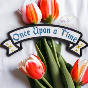 May include: White and red tulip flowers with a white banner with blue text that reads "Once Upon a Time" and two gold stars on each end of the banner.