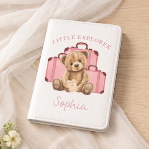 May include: White passport holder featuring a teddy bear illustration and pink suitcases. The text "Little Explorer" and the name "Sophia" are printed on the cover. The passport holder is made of a smooth, leather-like material.