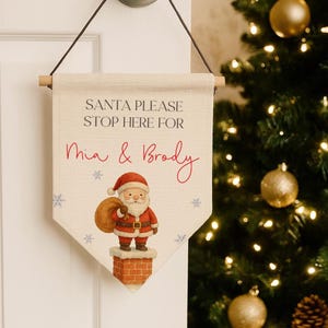 Op de afbeelding: Een kerstbanner met de tekst "SANTA PLEASE STOP HERE FOR Mia & Brody". Het toont een cartoon kerstman met een zak, staand op een schoorsteen, met sneeuwvlokken. De banner hangt aan een houten stok.